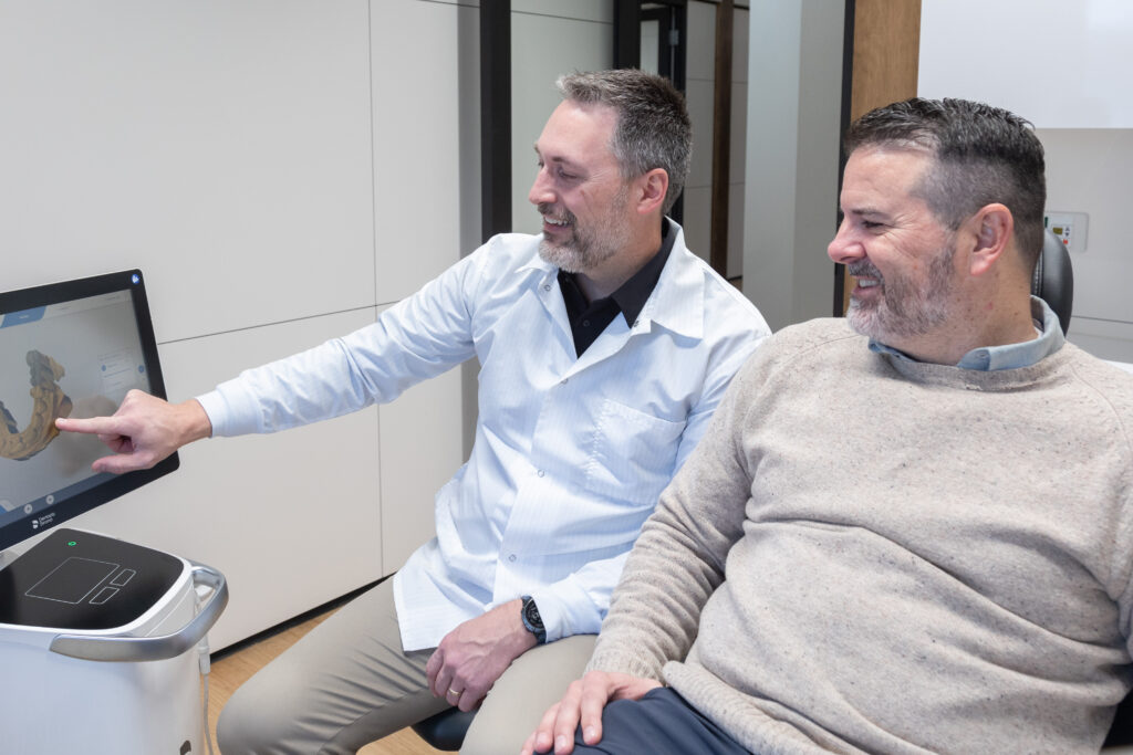 Dr. Conway in a white coat is smiling as he points to a 3D dental image on a computer screen. The patient, a middle-aged man with a beard wearing a beige sweater, is sitting in the dental chair next to the dentist, looking at the screen and smiling. They appear to be discussing the dental scan displayed on the monitor in a modern, well-equipped dental office.