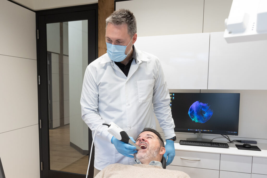 Dr. Conway of Spokane Valley Dentistry in Spokane Valley, WA, wearing a white coat, mask, and blue gloves uses an intraoral scanner to examine a patient's teeth. The patient, a middle-aged man with a beard, is reclining in the dental chair with his mouth open. Behind them, there is a computer monitor displaying a colorful 3D image of a dental scan. The dental office is modern with clean, white cabinetry and a large window providing natural light.