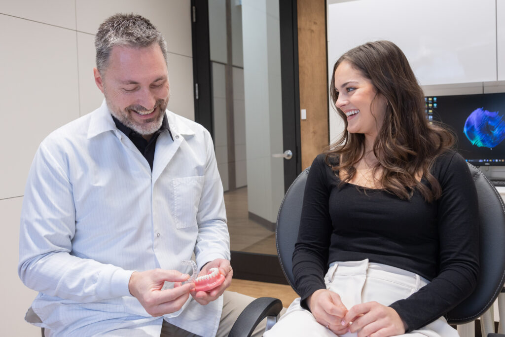 Dr. Conway in a white coat is smiling as he holds a dental model with a clear aligner. The patient, a young woman with long brown hair wearing a black top and white pants, is sitting in a dental chair next to him, looking at the model and smiling. They appear to be discussing the aligner and dental treatment in a modern, well-equipped dental office.