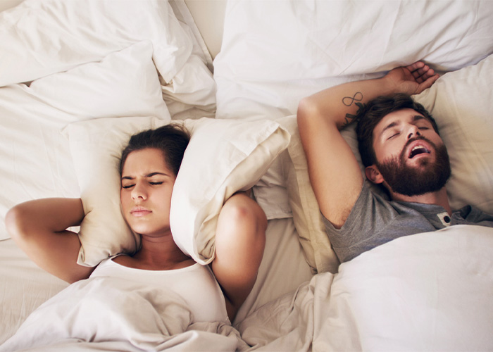 A couple is lying in bed with white sheets and pillows. The man on the right is sleeping with his mouth open and arms above his head. The woman on the left appears frustrated, covering her ears with a pillow to block out the noise. She has a pained expression on her face.
